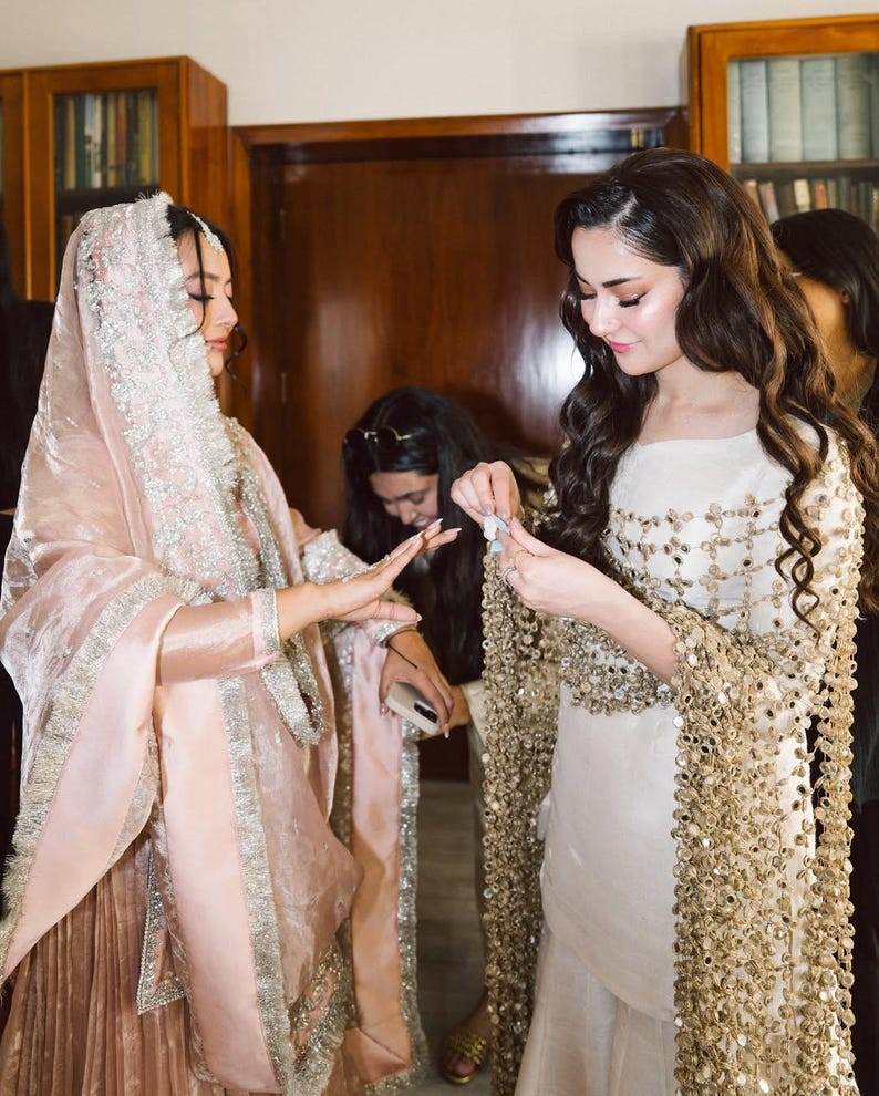 May include: Two individuals are shown in a room, one wearing a light pink outfit with silver trim and a matching head covering. The other is wearing a white dress with gold embellishments and a gold beaded shawl. The scene suggests a cultural or celebratory event.