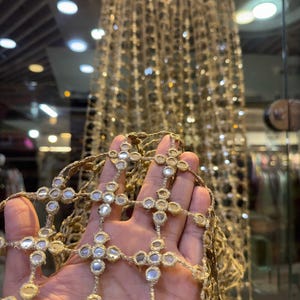 May include: Close-up of gold-colored decorative trim with reflective mirror accents. The trim is held in a hand, showcasing the intricate cross-like design. A larger piece of the same trim hangs in the background, suspended from a wooden hanger.