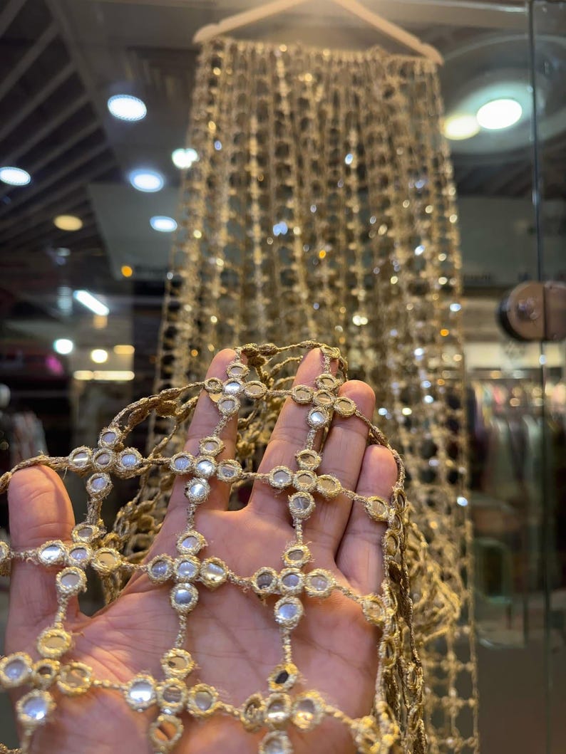 May include: Close-up of gold-colored decorative trim with reflective mirror accents. The trim is held in a hand, showcasing the intricate cross-like design. A larger piece of the same trim hangs in the background, suspended from a wooden hanger.