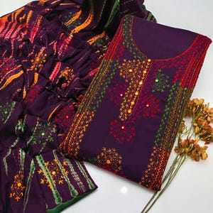 May include: A plum-colored embroidered salwar kameez set. The top features intricate embroidery in shades of orange, green, and red, with small mirror accents. The matching dupatta has a colorful striped pattern and embroidered details. A sprig of dried flowers is in the bottom right corner.