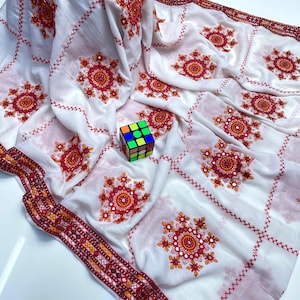 Op de afbeelding: Witte stof met een herhalend patroon van rode bloemenborduurwerk en kruissteekdetails. De stof heeft een decoratieve rand met een geometrisch ontwerp in rood, oranje en geel. Een Rubik's Cube ligt op de stof.