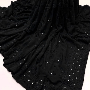 May include: A black fabric textile featuring intricate tonal embroidery and small circular mirror embellishments scattered throughout the material. The fabric is draped in soft folds, highlighting the detailed needlework and reflective accents.