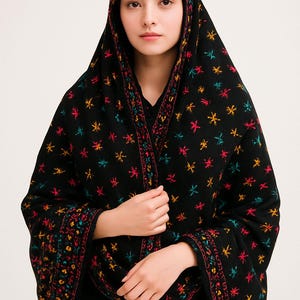 Puede incluir: Un chal negro con coloridos bordados florales. El chal está drapeado sobre la cabeza y los hombros, con los bordes con intrincados diseños en rojo, amarillo y azul. La prenda está hecha de una tela suave y fluida.