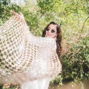 May include: A woman wearing a cream-colored, open-weave shawl with sparkling embellishments. She is smiling and wearing sunglasses and gold earrings. The background is a lush green forest with sunlight filtering through the trees.