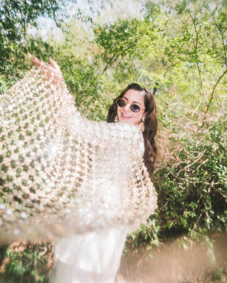 May include: A woman wearing a cream-colored, open-weave shawl with sparkling embellishments. She is smiling and wearing sunglasses and gold earrings. The background is a lush green forest with sunlight filtering through the trees.
