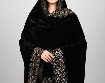 May include: A black velvet shawl with a hood and gold-coloured embellishments. The shawl drapes over the shoulders and features decorative trim along the edges and hood. The woman in the image is wearing the shawl.