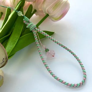 May include: A green and pink braided cord with a pink and green stone charm. The cord is tied in a knot at the top and has a small green bead at the end.