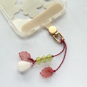 May include: Clear phone case with a beige grip and gold-colored hardware. A burgundy cord is attached to a charm with pink leaf-shaped beads, green beads, and a white teardrop-shaped bead. The case has a floral design.