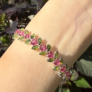 Pode incluir: Uma pulseira de prata com um design floral com pedras preciosas rosa e verdes. A pulseira é adornada com pedras rosa em forma de coração e pedras verdes em forma de folha. A pulseira está em um pulso.