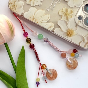 May include: Two beaded phone charms with colorful gemstones and a braided red and white cord. One charm has a circular, translucent pendant. A phone case with white floral designs is in the background.