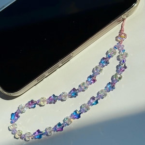 May include: A beaded phone charm with iridescent star and clear crystal beads. The charm is attached to a pink cord and is next to a smartphone.