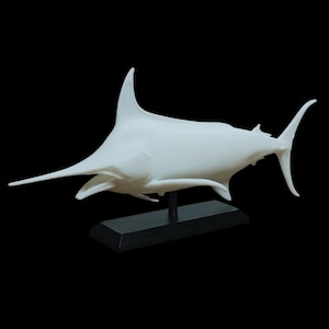 May include: A white marlin sculpture with a long, pointed bill and a tall dorsal fin. The sculpture is mounted on a black rectangular base, set against a black background. The marlin's streamlined body suggests speed and power.