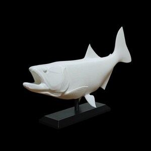 May include: A white sculpture of a fish with its mouth open, mounted on a black rectangular base. The fish has detailed fins and a streamlined body, set against a black background.