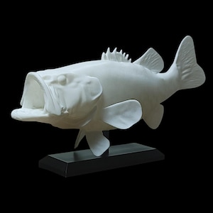 May include: A matte white sculpture of a largemouth bass fish with its mouth wide open, featuring detailed scales and fins. The fish is mounted on a rectangular black base, positioned horizontally to display its full body profile.