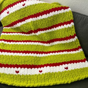 May include: A handmade, chunky knit blanket in a festive color scheme. The blanket features alternating stripes of lime green, red, and white. Small red hearts are scattered across the white stripes. The blanket is soft and cozy.