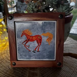 Può includere: Piastrella quadrata con cornice in legno raffigurante un cavallo rosso stilizzato con criniera e coda arancione su sfondo grigio. La cornice è marrone scuro con angoli decorativi in metallo. La piastrella è esposta su una superficie di legno.