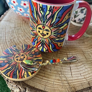 May include: A colorful ceramic mug with a sun design, a matching coaster, and a decorative spoon with colorful beads. The mug has a red interior and handle. The coaster and mug feature a vibrant sun face design.