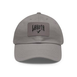 Peut inclure: Une casquette de baseball en coton gris avec un patch noir représentant le mot "LAKOTA" en police gothique avec un aigle noir.