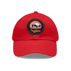 Peut inclure: Casquette de baseball rouge avec un patch noir et blanc représentant une silhouette de bison sur fond de coucher de soleil. Le patch porte le mot "Native" en lettres orange.