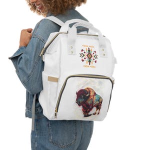 May include: A white backpack featuring a top handle and shoulder straps. The front displays a colorful geometric pattern above a large, detailed illustration of a bison with intricate, multi-colored markings and a textured, artistic appearance.