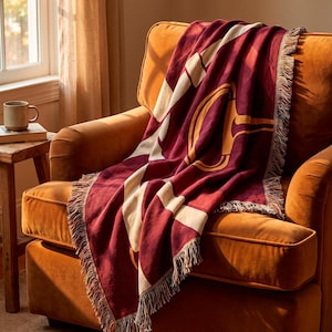 Op de afbeelding: Een geweven plaid in een dieprode en crèmekleurige kleurstelling met een groot, gestileerd goudkleurig bitmotief. De rechthoekige deken heeft een textuur en is afgewerkt met decoratieve franjes langs de randen.