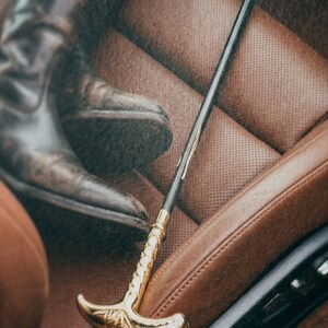 Puede incluir: Un bast&oacute;n negro y dorado con un mango curvo y detalles dorados decorativos. El bast&oacute;n est&aacute; colocado sobre un asiento de coche de cuero marr&oacute;n, con una bota de cuero marr&oacute;n en el fondo.