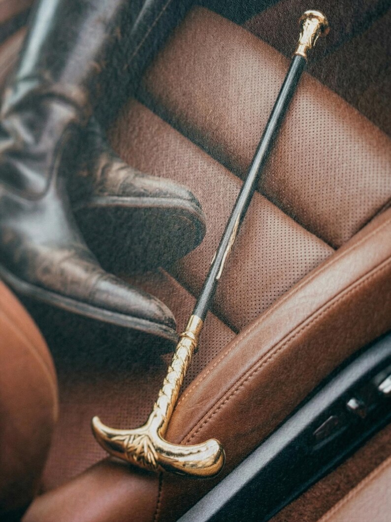 Puede incluir: Un bast&oacute;n negro y dorado con un mango curvo y detalles dorados decorativos. El bast&oacute;n est&aacute; colocado sobre un asiento de coche de cuero marr&oacute;n, con una bota de cuero marr&oacute;n en el fondo.