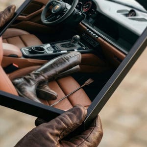 Puede incluir: Imagen enmarcada de un interior de coche con asientos de cuero marr&oacute;n, volante y palanca de cambios. Se ven unas botas de montar de cuero marr&oacute;n y una fusta. La persona que sostiene el marco lleva guantes de cuero marr&oacute;n.