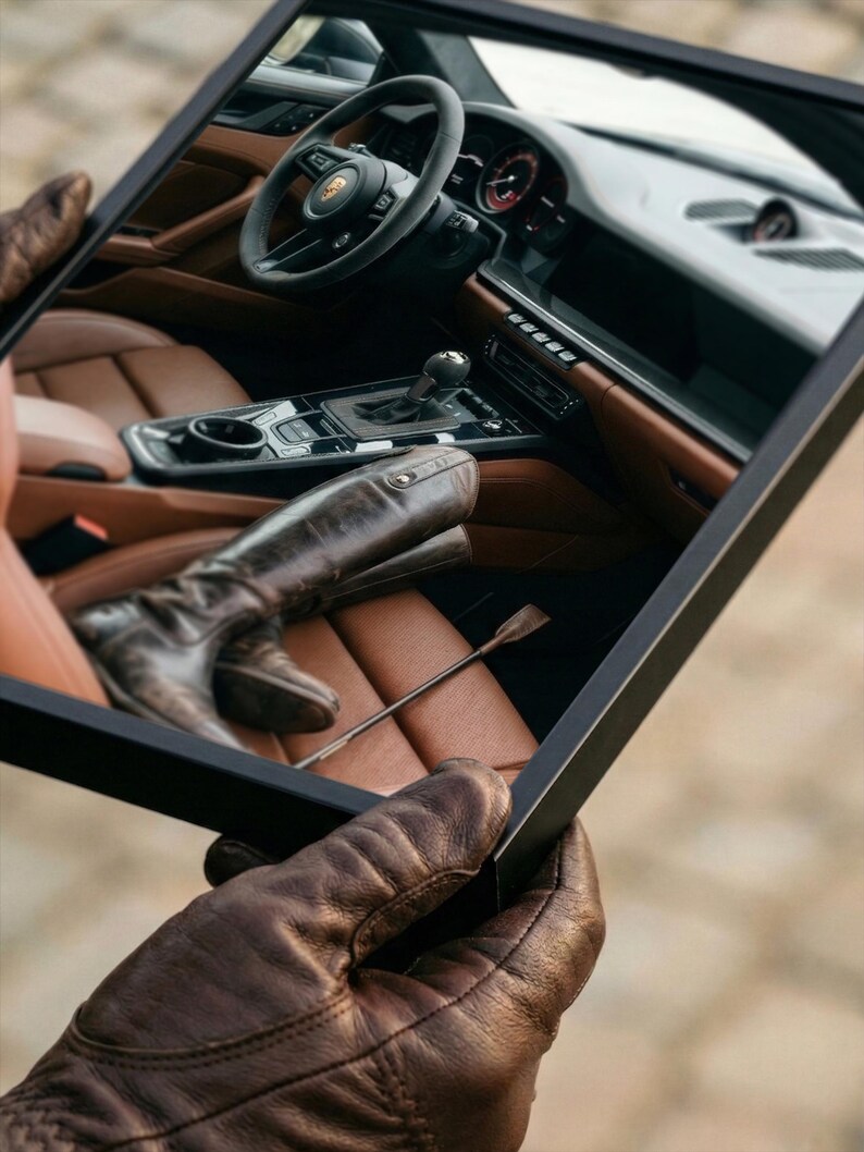 Puede incluir: Imagen enmarcada de un interior de coche con asientos de cuero marr&oacute;n, volante y palanca de cambios. Se ven unas botas de montar de cuero marr&oacute;n y una fusta. La persona que sostiene el marco lleva guantes de cuero marr&oacute;n.