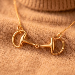 Peut inclure: Un collier doré avec un pendentif mors de cheval central. Le pendentif se compose de deux anneaux en forme de D reliés par une barre horizontale, suspendus à une fine chaîne dorée. Le métal présente une finition polie et réfléchissante.
