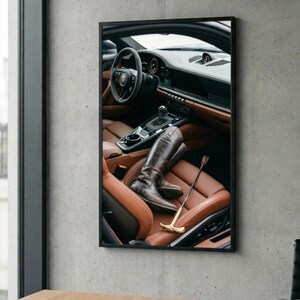 Puede incluir: Imagen enmarcada del interior de un coche con asientos de cuero marr&oacute;n. Un par de botas de montar marrones y una fusta con mango dorado descansan en el asiento. El volante y el salpicadero son visibles.