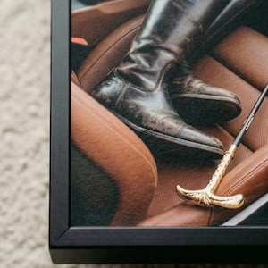 Puede incluir: Impresi&oacute;n enmarcada que muestra un par de botas de montar de cuero marr&oacute;n oscuro y desgastado sobre un asiento de coche de cuero marr&oacute;n. Tambi&eacute;n se ve un l&aacute;tigo de montar dorado. La imagen est&aacute; enmarcada en un marco negro.
