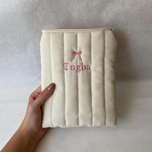 Puede incluir: Un estuche con cremallera de color crema, acolchado verticalmente, con un lazo rosa bordado y el nombre "Tugba" en letras rosas centrado en la parte frontal. El estuche tiene un acabado de tela suave y texturizada y una cremallera color crema.