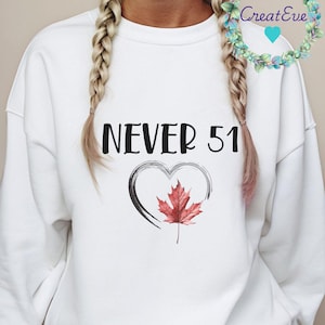 May include: White sweatshirt with the text "NEVER 51" in black. A black heart outline surrounds a red maple leaf. The sweatshirt is a casual style.