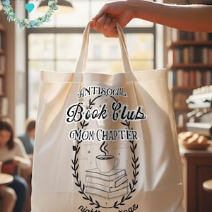 Op de afbeelding: Cr&egrave;mekleurige draagtas met de tekst "Antisocial Book Club Mom Chapter nightly meetings" in zwart schrift. Het ontwerp bevat een koffiekopje bovenop een stapel boeken, omringd door een lauwerkrans. De tas wordt vastgehouden.
