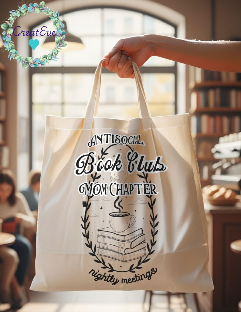 Op de afbeelding: Cr&egrave;mekleurige draagtas met de tekst "Antisocial Book Club Mom Chapter nightly meetings" in zwart schrift. Het ontwerp bevat een koffiekopje bovenop een stapel boeken, omringd door een lauwerkrans. De tas wordt vastgehouden.