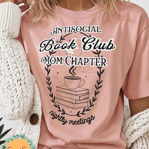 Op de afbeelding: Lichtroze t-shirt met de tekst "Antisocial Book Club Mom Chapter" in een decoratief lettertype. Het ontwerp bevat een stapel boeken, een koffiekopje en een krans. De zin "nightly meetings" is ook opgenomen.