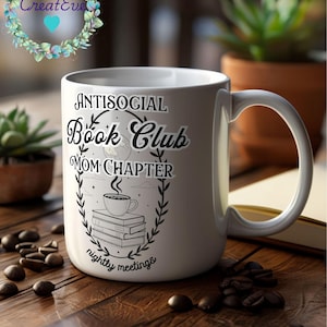Op de afbeelding: Witte keramische mok met de tekst "ANTISOCIAL Book Club MOM CHAPTER nightly meetings" in zwart. Het ontwerp bevat een koffiekopje, boeken en een maan. De mok staat op een houten oppervlak met koffiebonen.