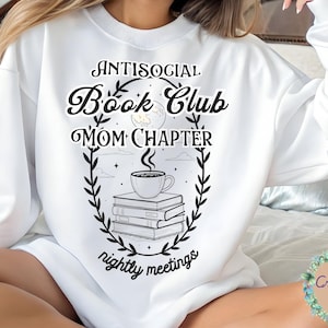 Op de afbeelding: Witte sweatshirt met de tekst "Antisocial Book Club Mom Chapter nightly meetings" in zwarte, decoratieve letters. Een afbeelding van een koffiekopje op boeken staat in het midden, omlijst door een krans.