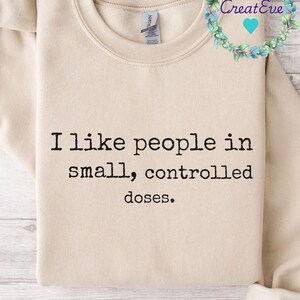 Op de afbeelding: Een beige sweatshirt met de tekst "Ik hou van mensen in kleine, gecontroleerde doses." in het zwart gedrukt.
