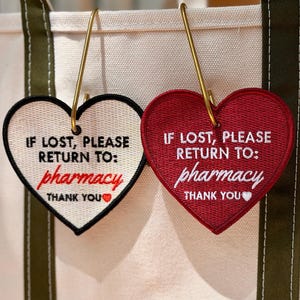 May include: Two heart-shaped embroidered bag tags hanging from a canvas tote. One tag is cream with black text and a red heart, the other is red with white text and a white heart. Both read: IF LOST, PLEASE RETURN TO: pharmacy THANK YOU.