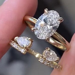 Pode incluir: Um anel de noivado de ouro com um grande diamante oval. O anel é adornado com diamantes menores na banda. O anel é usado em um dedo com outras duas alianças de ouro com diamantes em forma de pêra.