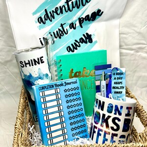 May include: A wicker basket filled with reading-themed gifts. The items include a blue and white tumbler with the word "SHINE" on it, a blue journal with the words "COMPLETION BOOK JOURNAL" and a star rating system, a teal journal with the words "take a chance" on the cover, a blue and white mug with the words "IN ON BOOKS COFFEE" on it, a blue bookmark with the words "A BOOK A DAY KEEPS REALITY AWAY" on it, and a white tote bag with the words "adventure is just a page away" on it.