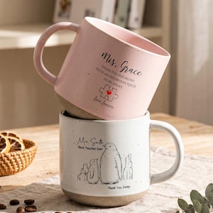 May include: Two ceramic mugs stacked vertically. The top mug is light pink with a speckled finish, featuring the text "Mis. Grace" and a puzzle piece graphic. The bottom mug is white with a speckled finish, featuring a penguin illustration and the text "Mrs. Smith Best Teacher Ever". Both mugs have a natural, unglazed tan base.