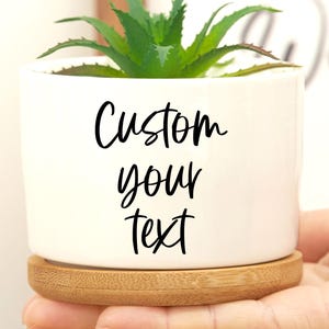 May include: A white ceramic planter with a green succulent plant. The planter has the words "Custom your text" in black script. The planter sits on a round, natural wood saucer.