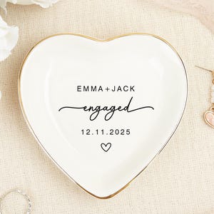 Peut inclure: Un vide-poches blanc en forme de cœur avec un bord doré, portant l'inscription "EMMA + JACK engaged 12.11.2025". Deux paires de boucles d'oreilles avec des pendentifs en forme de cœur rose et une bague en argent sont visibles.