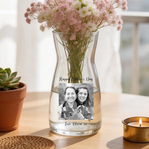 May include: A clear glass vase with a photo and text that reads "Happy Mother's Day" and "Love Olivia xx". The vase holds pink and white flowers. A small succulent in a terracotta pot, a woven coaster, and a lit candle in a gold holder are also on the table.