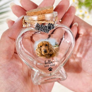 May include: A heart-shaped glass keepsake with a cork stopper, featuring a photo of a dog. The jar is labeled "Dog Person" and "Bella 2003-2023". A paw print design is at the bottom. The jar is held in two hands.