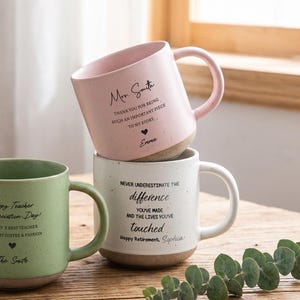 May include: Three ceramic mugs in light pink, sage green, and white with speckled textures and dark text. A clear 10 cm ruler stands next to the green mug. The mugs feature personalised messages and names printed in black, with a handle on each side.