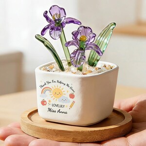 May include: A small white square ceramic planter containing two purple glass flowers with green stems and clear pebbles. The planter features a printed graphic of a sun, rainbow, clouds, apples, and a pencil with the text Thank You For Helping Me Grow, LOVE, LILY, Miss Anna.